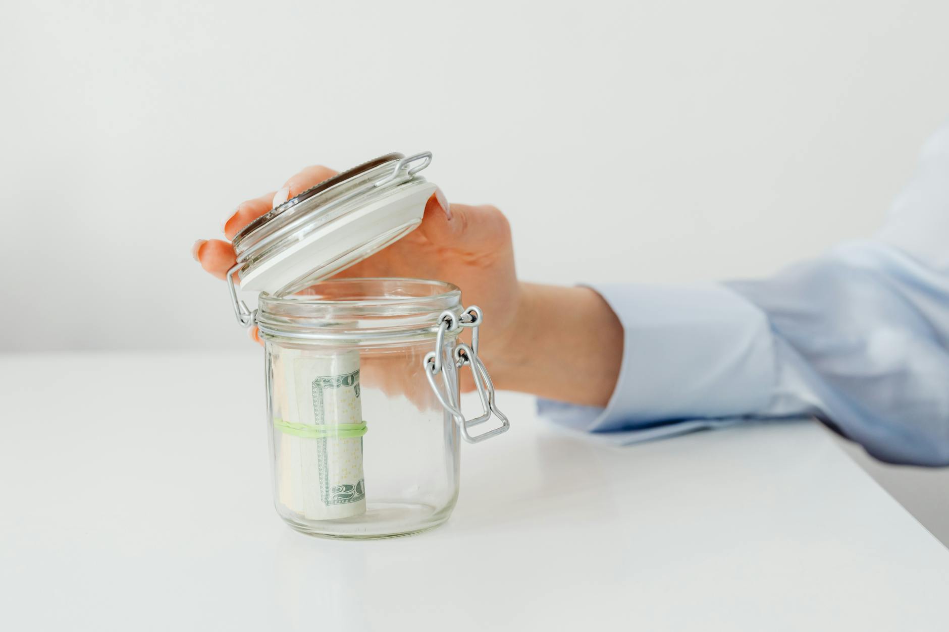 A hand lifting the lid of a glass jar holding a roll of dollar bills on a white table