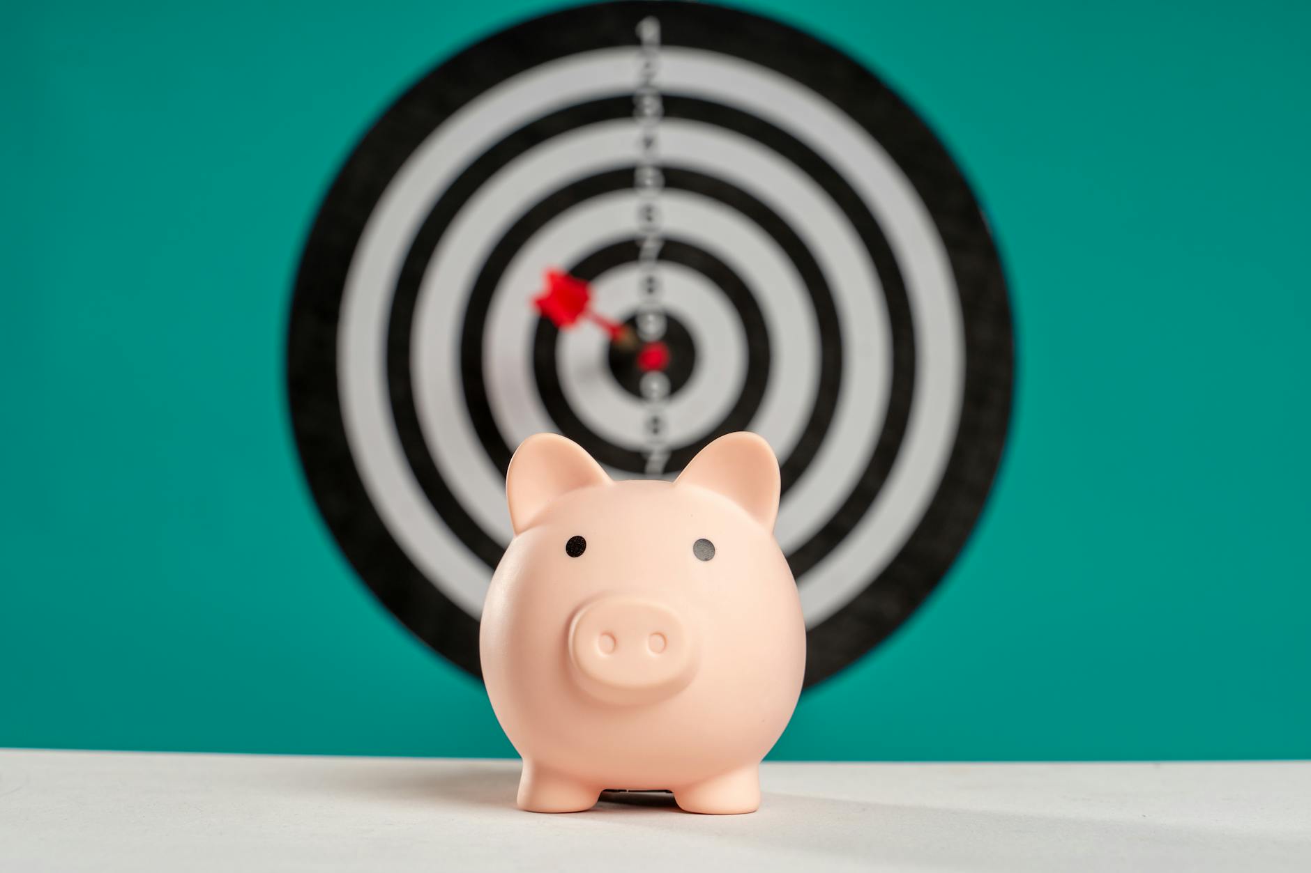 A pink piggy bank in front of a dartboard with a dart near the center