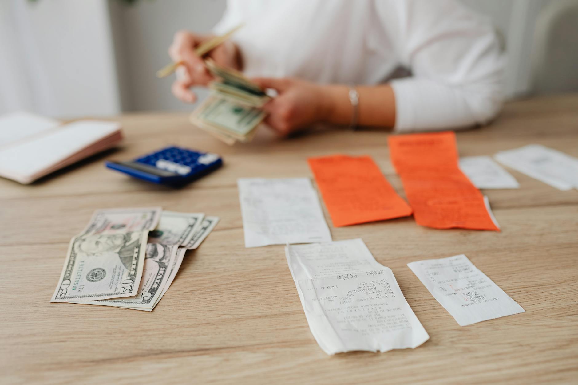 A hand holding dollar bills over a desk with receipts, cash, and a calculator