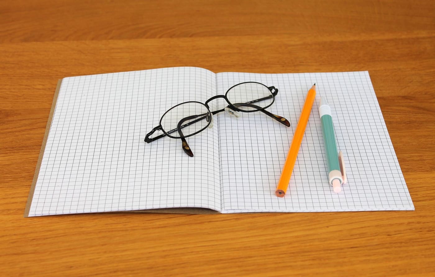 An open graph-paper notebook with glasses and two pencils on a wooden table