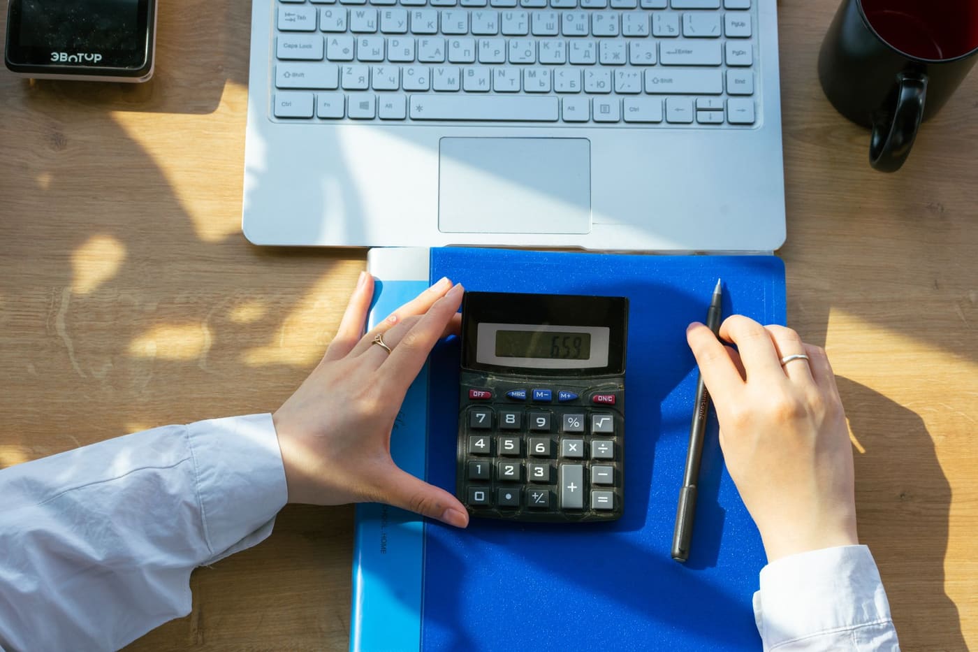 Hands using a desktop calculator next to a laptop, a pen, and a notebook