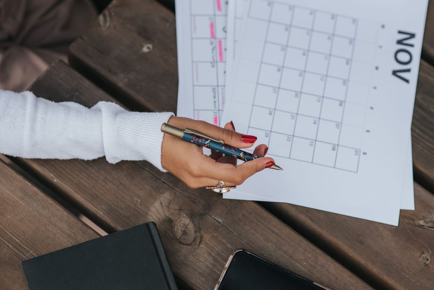 A hand holding a pen over a printed monthly calendar on a wooden table